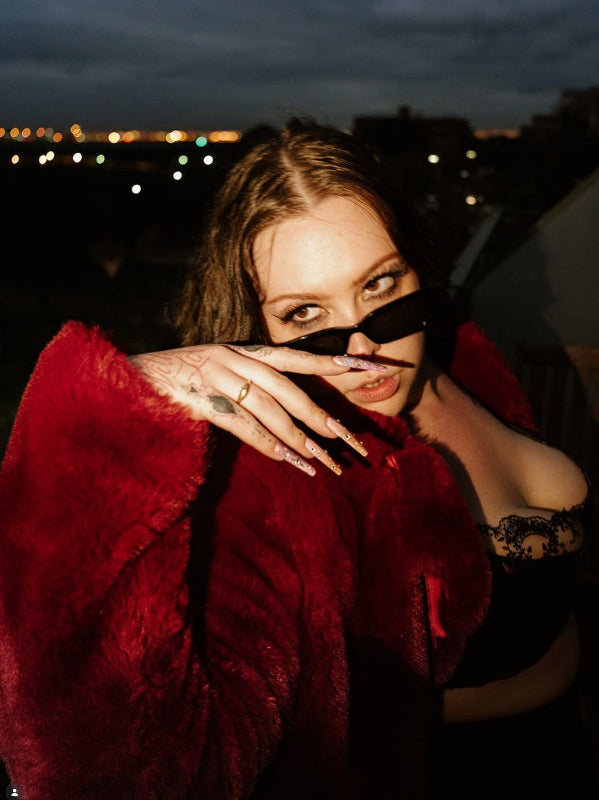 A stylish woman with bold makeup and long manicured nails, wearing a red faux fur coat and black lingerie, poses outdoors at night with city lights in the background, highlighting her confident attitude.