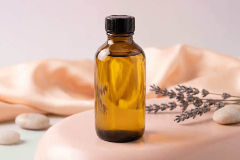 Brown glass bottle with black cap on a light pink fabric background with lavender and stones. Bulk refill 100ml cuticle oil for wholesale australian nail srerum.