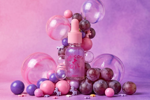 A pink dropper bottle labeled 'Unicorn Oil' surrounded by pink, purple, and clear decorative bubbles and glossy spheres on a pink-purple studio background.