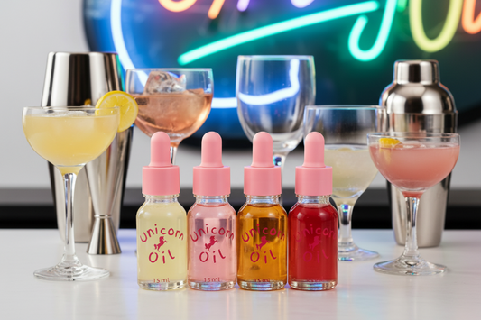 Four bottles of 'Unicorn Oil' with colorful liquids on a bar counter with cocktails and neon lights in the background.