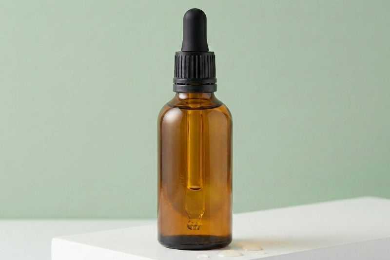 Amber glass dropper bottle with black cap on a white surface and light green background. bulk wholesale cuticle oil refill