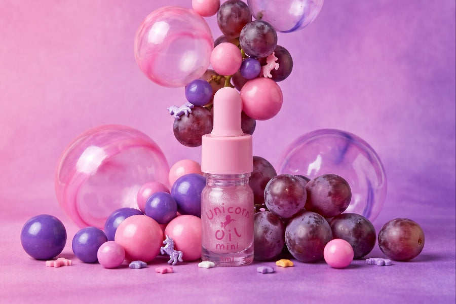 A pink dropper bottle labeled 'unicorn oil mini' styled with pink and purple grapes, translucent balloons, and pastel beads on a pink and purple gradient background.