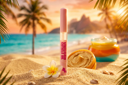 A pink cosmetic tube labeled 'UNICORN CUTICLE OIL' stands on sandy beach ground with a tropical ocean and palm trees in the background. A rolled towel, a flower, and a jar of colorful cream are also visible in the scene.