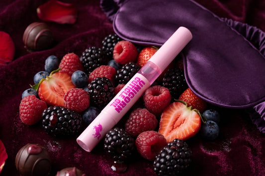 A pink cuticle oil pen labeled 'Blossom' lies on a bed of mixed berries, chocolate-covered chocolates, red rose petals, and a purple satin sleep mask on a deep burgundy velvet fabric background.