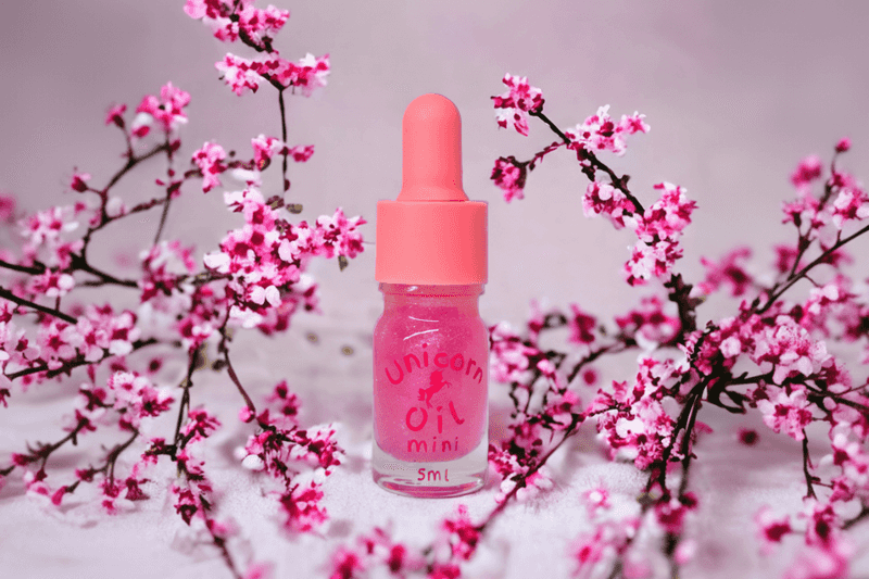 Pink cuticle oil dropper bottle labeled 'Unicorn Oil' with cherry blossoms in the background