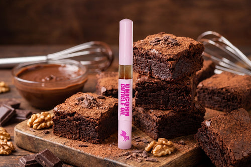 A tube of pink cuticle oil labeled 'UNICORN CAPSULE OIL' stands upright in front of stacked chocolate brownies on a wooden board, with chocolate, walnuts, and whiskers in the background.