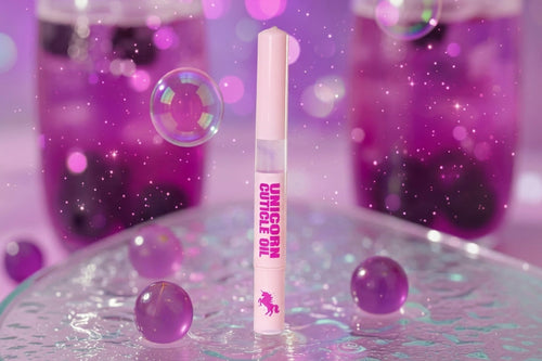 A single tube of unicorn cuticle oil stands upright on a reflective glass surface, with pink and purple bubbles and sparkling bokeh in the background.