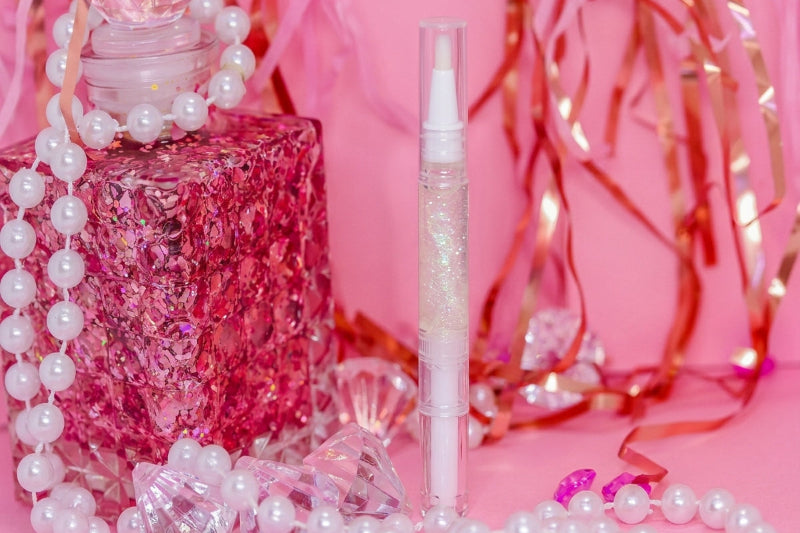 A clear tube of cuticle oil stands upright in the center, surrounded by pink decorative props including a pink glittery perfume bottle, white pearl beads, and shiny red ribbon against a pink background.