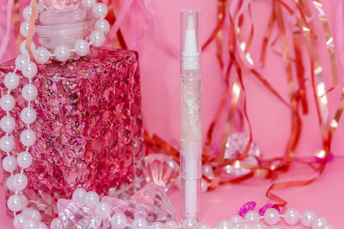 A clear tube of cuticle oil stands upright in the center, surrounded by pink decorative props including a pink glittery perfume bottle, white pearl beads, and shiny red ribbon against a pink background.