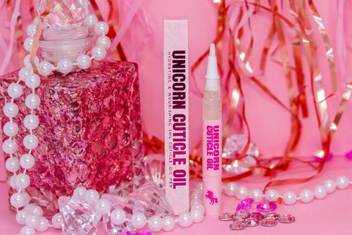 Unicorn Cuticle Oil set with a pink glittery bottle, a white tube, and a white box, styled with white pearl beads, pink tinsel, and pink gems on a pink background.
