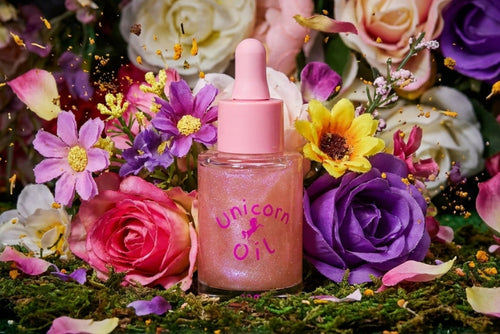 A pink dropper bottle labeled 'Unicorn Oil' surrounded by colorful flowers and green moss in a styled floral arrangement.