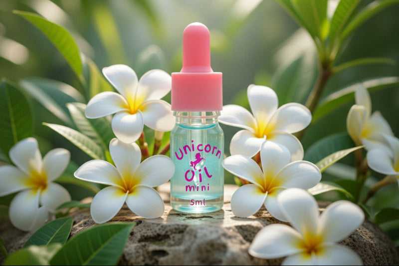 Bottle of Unicorn Oil Mini with a pink cap on a stone surface surrounded by white flowers.