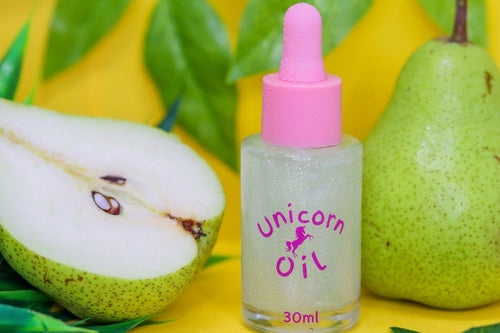 A 30ml bottle of 'Unicorn Oil' with a pink dropper cap, set against a bright yellow background with green pears and leaves. The bottle contains a sparkly, pear-shaped clear oil and features a pink label with a unicorn graphic.