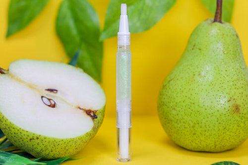 A clear cosmetic tube with a white applicator stands upright on a bright yellow surface, styled with a whole pear, a cut pear showing white flesh, and green leaves.