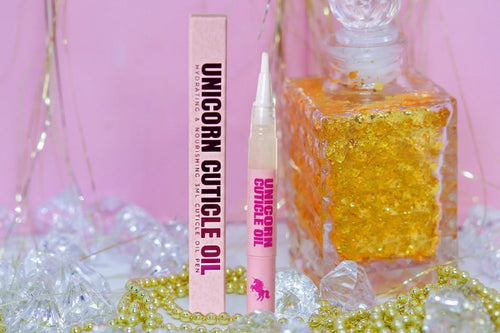 Unicorn Cuticle Oil set with a pink box, applicator tube, and a decorative clear glass bottle filled with gold glitter, styled with pearls and crystals on a pink background.