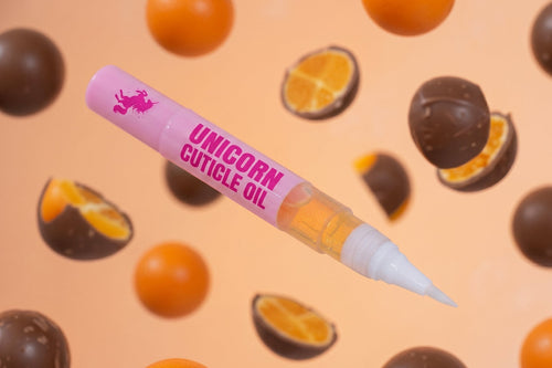 A pink applicator tube labeled 'UNICORN CUTICLE OIL' floating against a peach background with floating orange slices and chocolate-covered spheres.