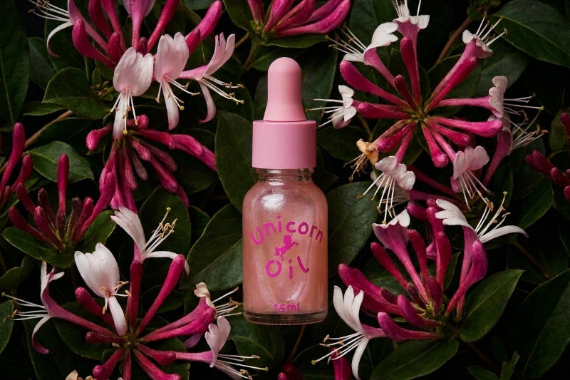 A small glass dropper bottle labeled 'Unicorn Oil' with a pink cap, centered among pink and white flowers and green leaves. The product appears to be a cosmetic or skincare oil, photographed in a styled flat-lay arrangement.