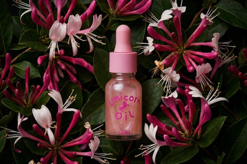 A small glass dropper bottle labeled 'Unicorn Oil' with a pink cap, centered among pink and white flowers and green leaves. The product appears to be a cosmetic or skincare oil, photographed in a styled flat-lay arrangement.