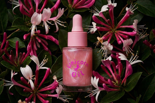A pink dropper bottle labeled 'Unicorn Oil' placed among pink and white flowers with green leaves, creating a natural floral background.