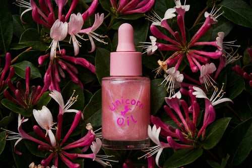 A pink dropper bottle labeled 'Unicorn Oil' placed among pink and white flowers with green leaves, creating a natural floral background.