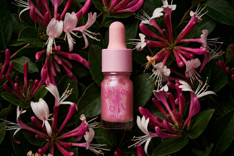 A pink dropper bottle labeled 'Unicorn Oil Mini' centered among dark green leaves and pink-and-white flowers.