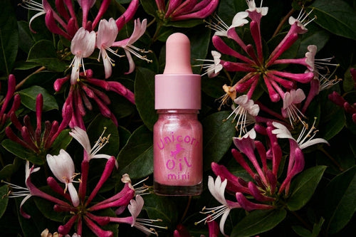 A pink dropper bottle labeled 'Unicorn Oil Mini' centered among dark green leaves and pink-and-white flowers.