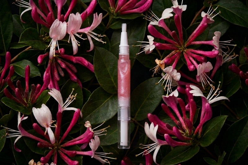 A cuticle oil pen with a pink heart-shaped cap and pink gloss inside, placed vertically on a bed of dark green leaves and magenta-pink honeysuckle flowers.
