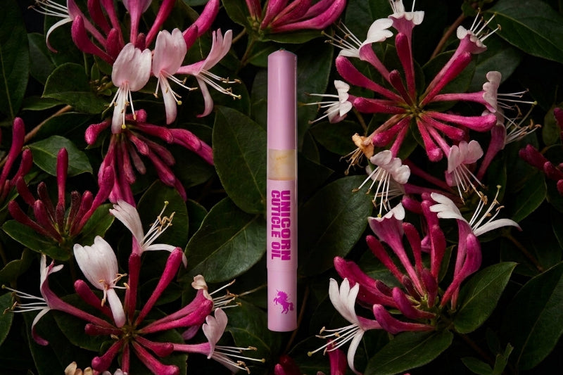 A pink cosmetic tube labeled 'UNICORN CUTICLE OIL' stands upright in the center against a background of dark green leaves and pink-and-white flowers.