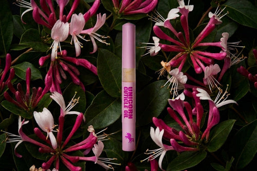 A pink cosmetic tube labeled 'UNICORN CUTICLE OIL' stands upright in the center against a background of dark green leaves and pink-and-white flowers.