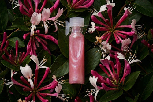 A clear plastic bottle filled with pink liquid and topped with a white cap, centered among dark green leaves and pink-and-white flowers.