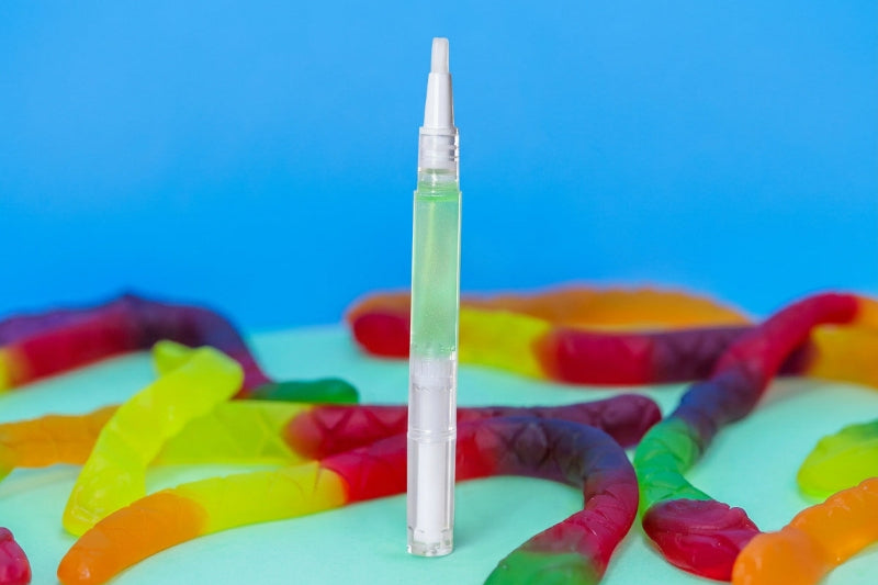 A clear plastic cuticle pen stands upright on a mint green surface, surrounded by colorful gummy worms, against a bright blue background.