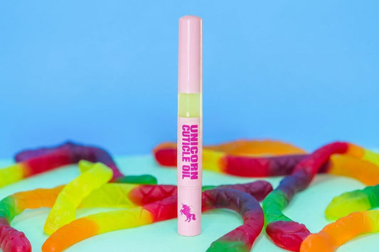 A pink tube labeled 'UNICORN CUTICLE OIL' stands upright on a white surface scattered with rainbow gummy worms, against a bright blue background.