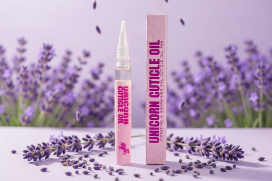 A tube of unicorn cuticle oil and its matching pink box are displayed on a white surface with scattered lavender buds and sprigs, against a soft purple lavender field background.