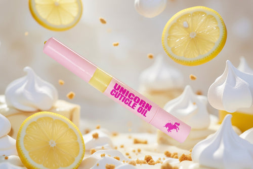 A pink cosmetic tube labeled 'UNICORN CUTICLE OIL' with a small unicorn logo, surrounded by floating lemon slices, whipped cream, and crumbs in a bright, airy studio setup.