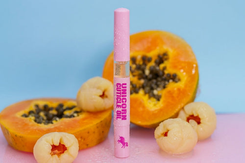 A pink cosmetic tube labeled 'UNICORN CUTICLE OIL' stands upright between sliced papayas and peeled fruit pieces on a pink surface against a light blue background.