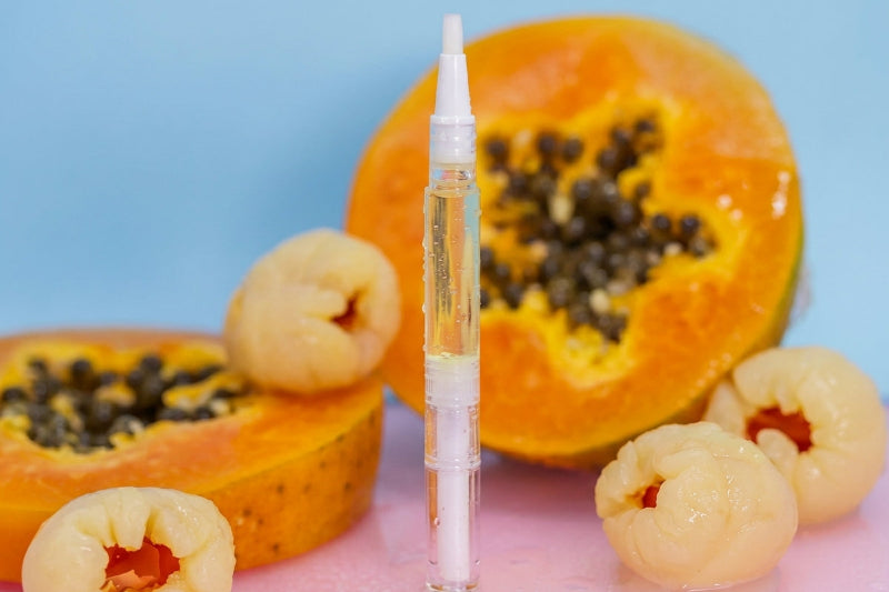 A clear cuticle oil pen with a white cap stands upright in the center, surrounded by sliced papayas and lychee fruits on a pink surface against a light blue background.