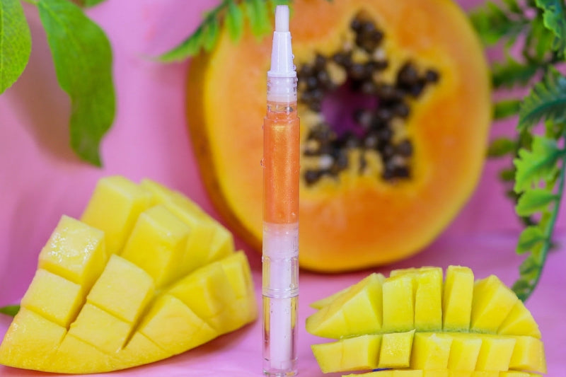 A clear cuticle oil pen with orange and white layers stands in the center, surrounded by sliced yellow mango and a halved papaya on a pink background with green leaves.
