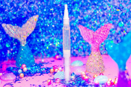 A clear cosmetic pen-style tube with a white applicator cap stands upright on a pink glitter surface, surrounded by pastel pearls and mermaid tail decorations against a sparkling blue background.