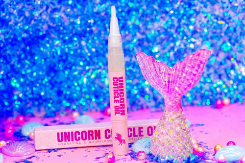 A pink cosmetic tube labeled 'UNICORN OIL' stands upright next to its matching pink box, with a glittery mermaid tail prop and pastel pearls on a pink surface against a sparkling blue background.