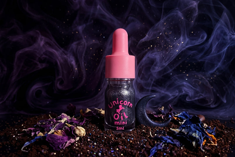 Black shimmer Unicorn Oil bottle with pink dropper on black marble, styled with coffee beans, vanilla pods, white flowers, warm candlelight, and smoky midnight accents.