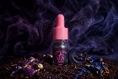 Black shimmer Unicorn Oil bottle with pink dropper on black marble, styled with coffee beans, vanilla pods, white flowers, warm candlelight, and smoky midnight accents.