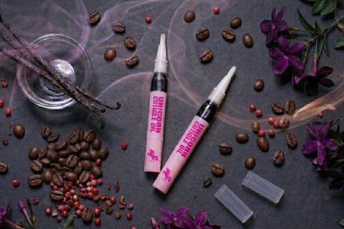 Two pink and black cosmetic tubes labeled 'UNIQUELY CHILEAN' and 'CHILEAN GEL' are arranged on a dark textured surface with scattered coffee beans, purple flowers, vanilla beans, and smoky decorative elements.