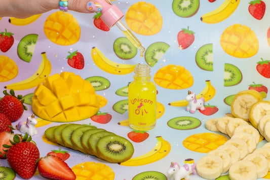 A hand holds a pink dropper above a small bottle of yellow liquid on a colorful fruit-patterned surface, surrounded by strawberries, kiwi slices, bananas, and sliced mangoes.
