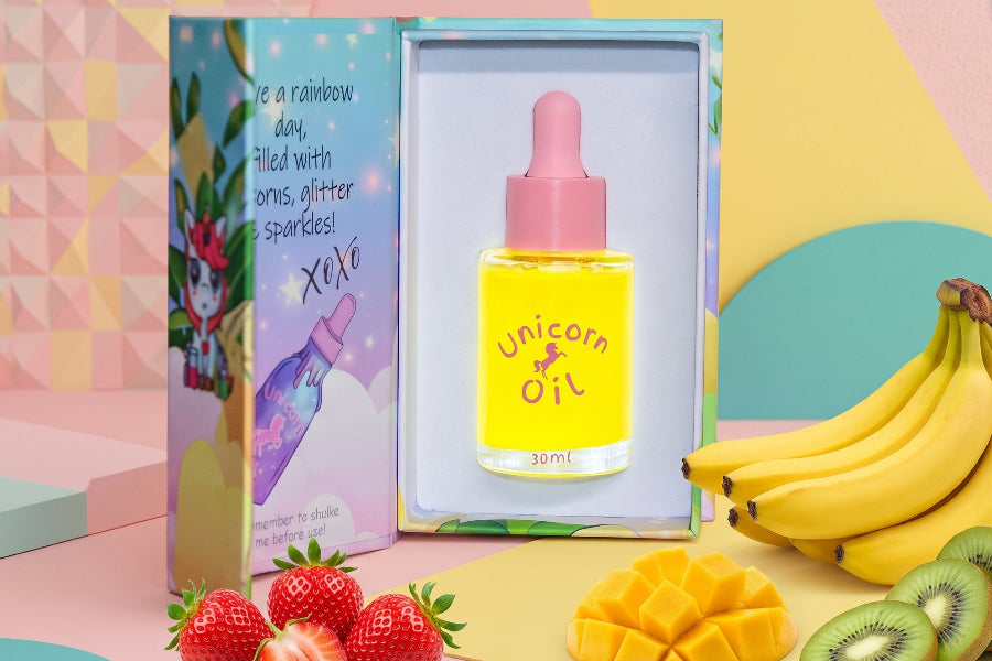 A 30ml bottle of Unicorn Oil displayed in its open packaging, surrounded by colorful props including strawberries, kiwi, mango, and bananas on a pastel background.