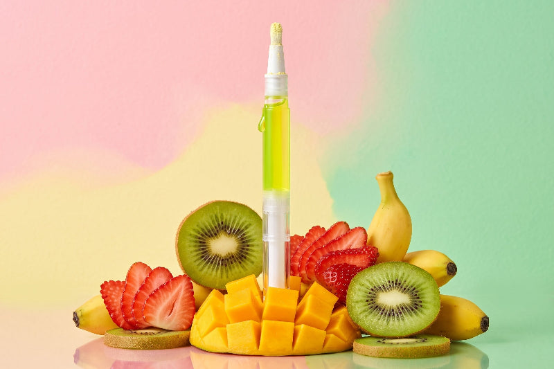 A single clear and white dropper-style bottle filled with bright yellow-green liquid stands upright in the center, surrounded by sliced kiwi, strawberries, mango cubes, and bananas on a pastel pink, yellow, and mint green background.
