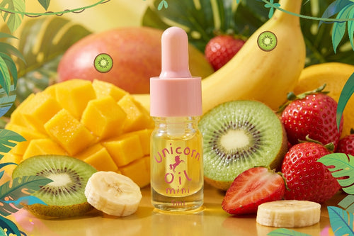 A small clear bottle of unicorn oil with a pink dropper cap stands centered among tropical fruits including mango, kiwi, strawberries, and banana slices on a bright yellow surface.