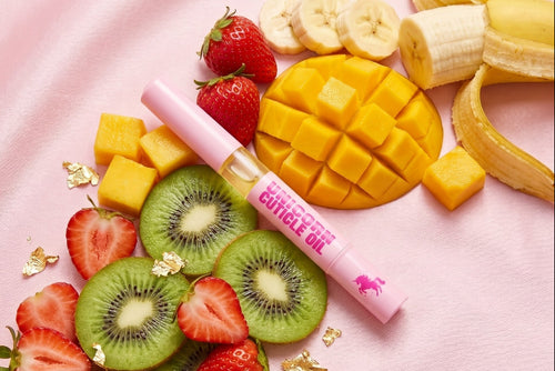 Pink cuticle oil tube labeled 'CUTICLE OIL' placed on a pink fabric background with sliced strawberries, kiwi, banana, mango, and gold flakes around it.