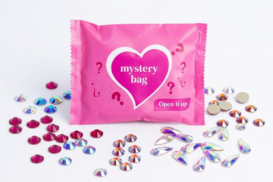 A pink mystery bag with a white heart graphic and the words 'mystery bag' and 'Open it up' on the front, surrounded by scattered iridescent rhinestones and small round beads on a white background.