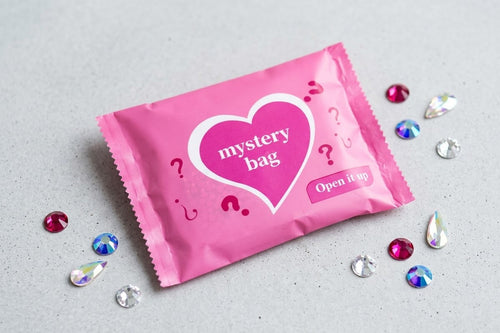 A pink foil mystery bag with a white heart graphic and the words 'mystery bag' and 'Open if possible' lies on a light gray surface, surrounded by scattered rhinestones.