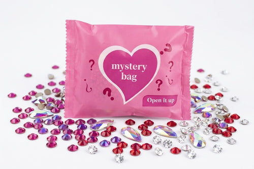 A pink foil mystery bag with a heart-shaped logo and the words 'mystery bag' and 'Open it up' sits on a white background, surrounded by scattered red, pink, silver, and iridescent rhinestones.